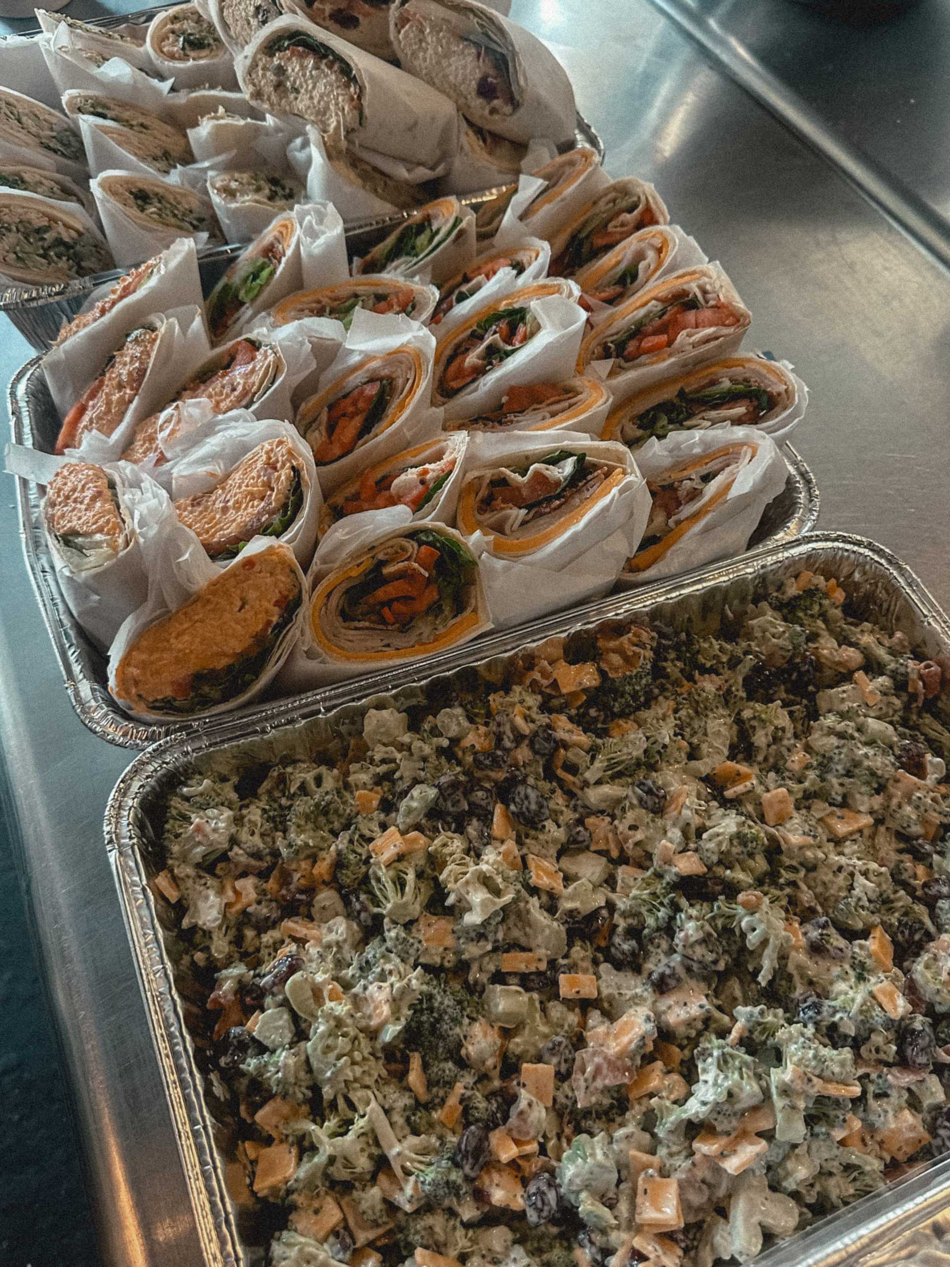 Deli wraps and trays of creamy broccoli salad on a metal counter.