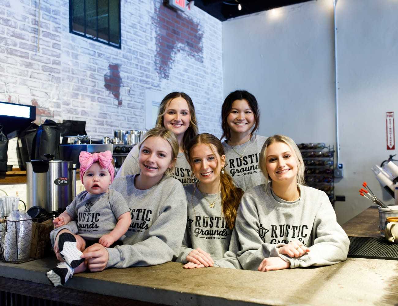 Family posing behind coffee shop counter in matching "Rustic Grounds" sweatshirts.