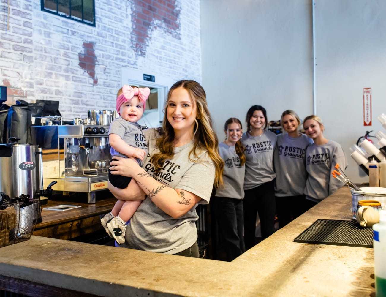 Staff posing together at a coffee shop counter, woman in front holding a baby.